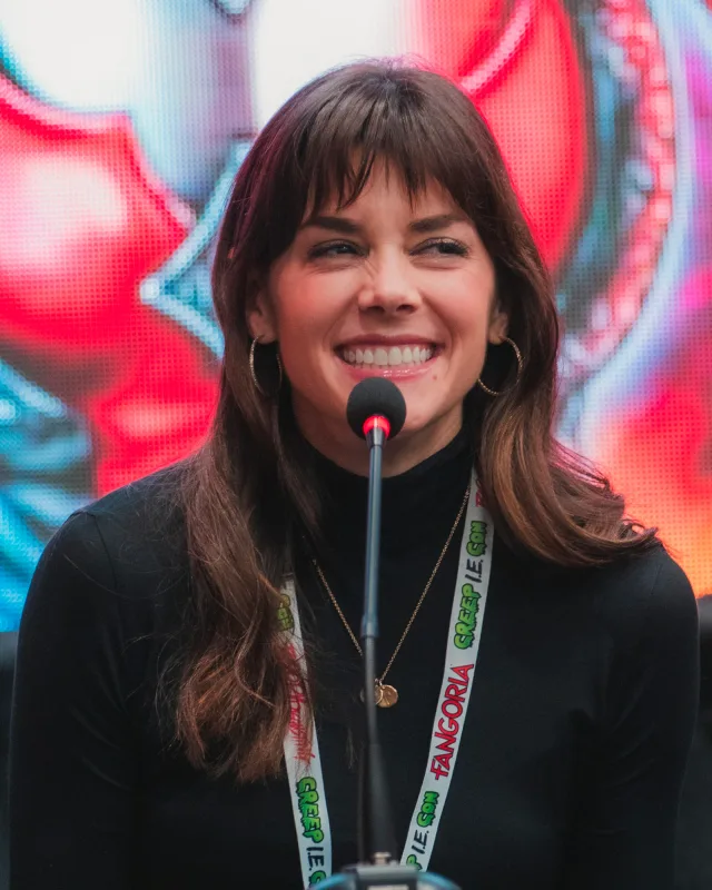 A woman with brown hair and a fringe, in a black polo neck and a thin gold necklace, laughing as she speaks into a microphone at a convention.