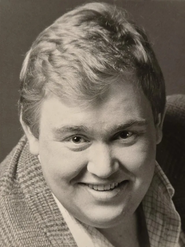 Black and white studio portrait of a smiling man with short fair hair, wearing a tweed jacket over a checked shirt.
