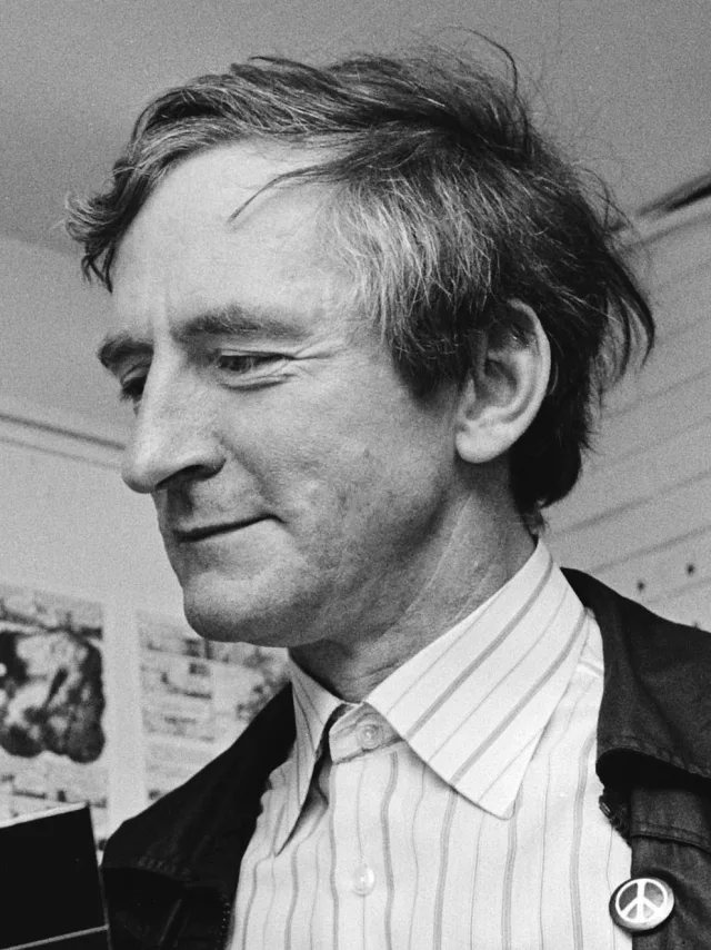 Black and white close portrait of Raymond Briggs in profile, tousled hair, striped shirt and dark jacket with a peace badge, looking down.