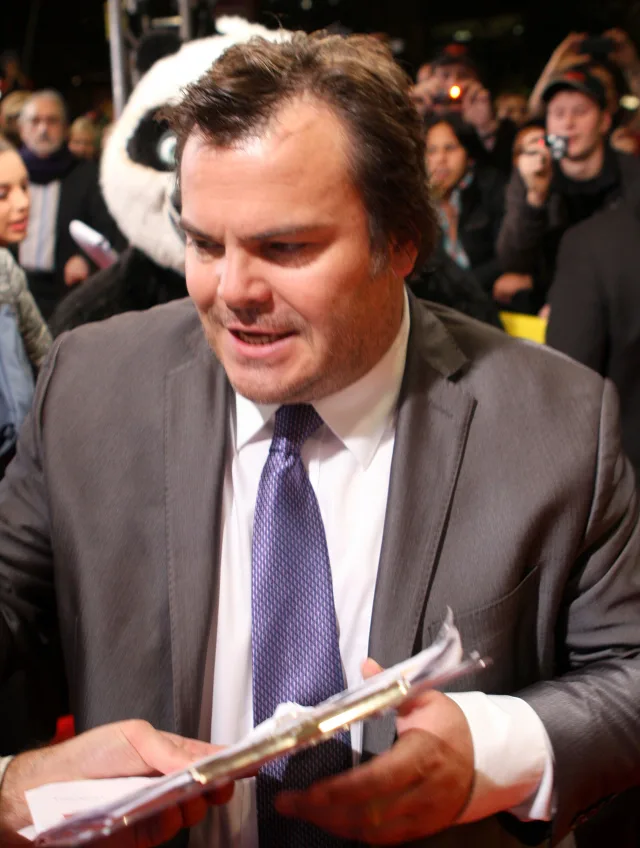 Head and shoulders photograph of a man with brown hair in a grey jacket and purple tie, signing autographs outdoors at an evening event.