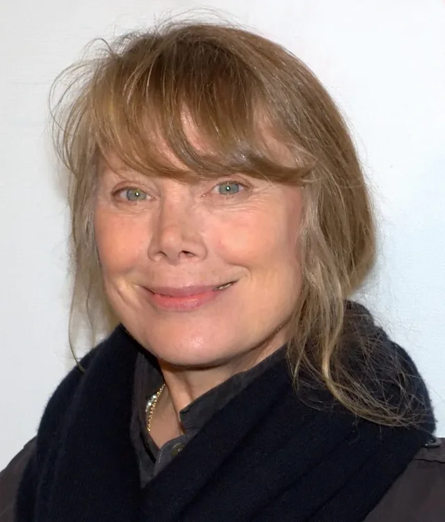 Smiling woman with fair, tousled hair and a fringe, wearing a dark scarf and jacket against a plain white background.