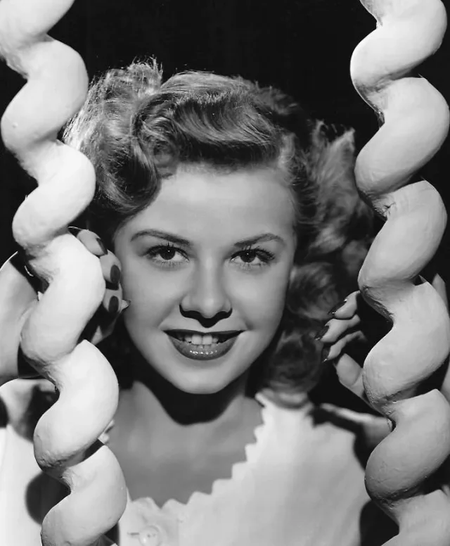 Black and white studio portrait of a smiling young woman with wavy hair, framed between two curling sculpted shapes.