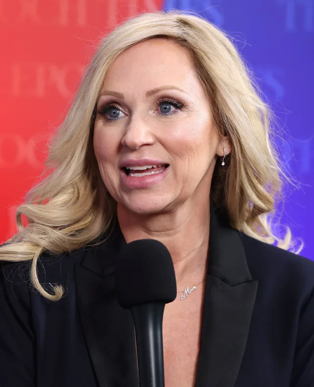Close photograph of a smiling blonde woman with wavy hair and a dark blazer speaking into a microphone against a red and blue backdrop.