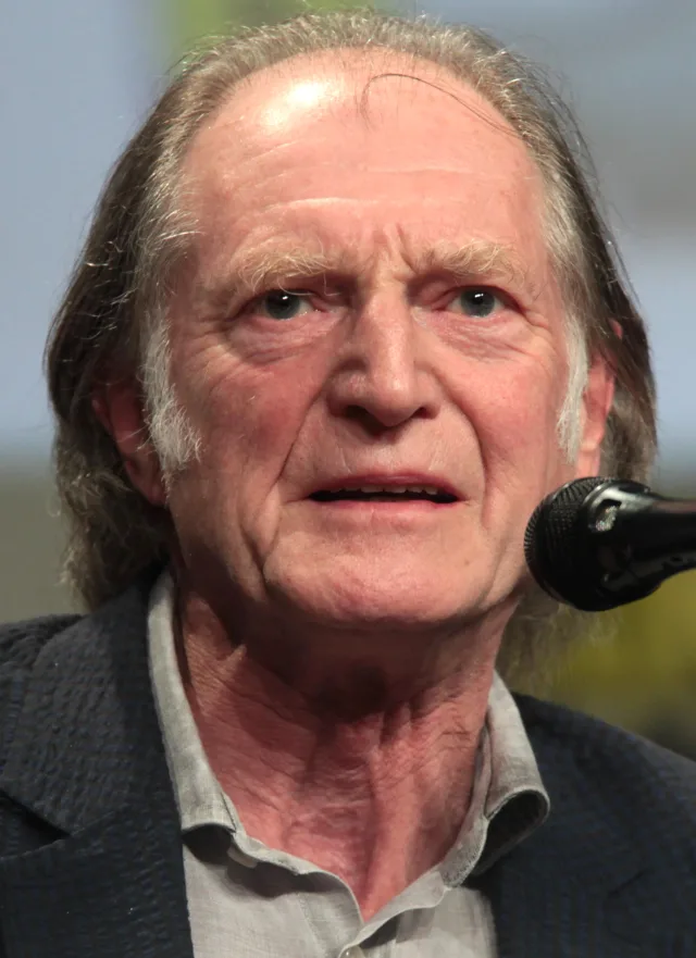 Close portrait of an older man with long grey hair and a navy jacket speaking beside a microphone at a convention panel.