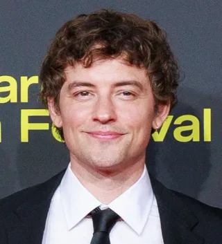 Head and shoulders photograph of a man with curly brown hair and a slight smile, in a white shirt, black tie and dark jacket.