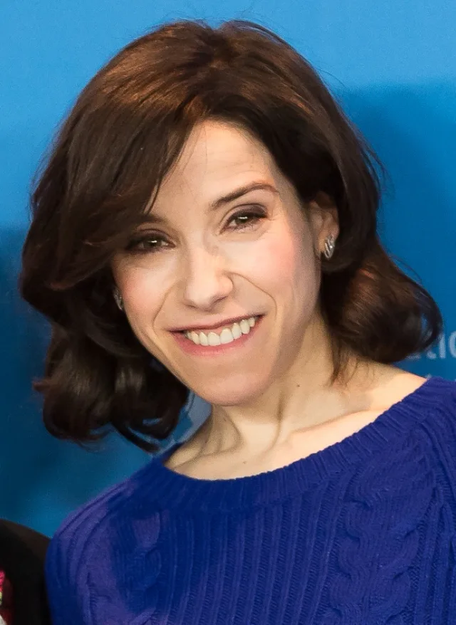 Smiling woman with shoulder-length dark brown hair in a royal blue knitted jumper, against a blue backdrop.