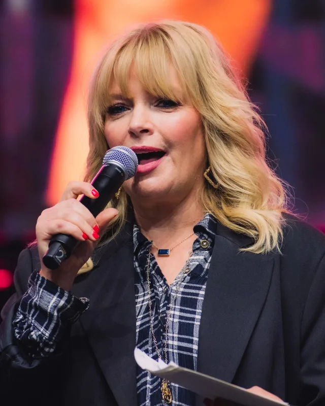 Woman with wavy blonde hair and fringe speaking into a microphone, wearing a dark blazer over a checked shirt.