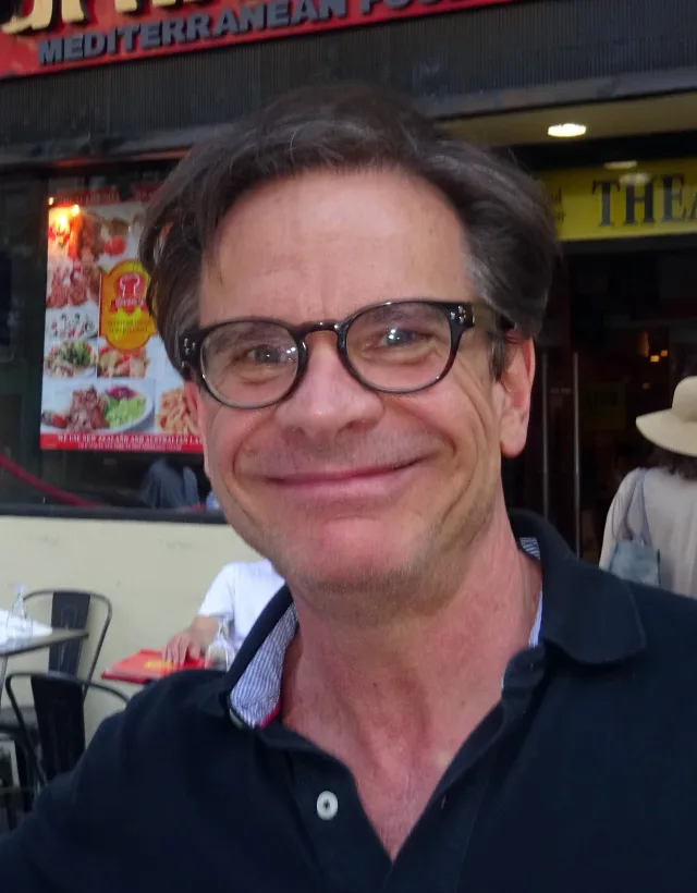 Smiling man with dark swept hair and thick dark-framed glasses, wearing a navy polo shirt, photographed outdoors at a street cafe.