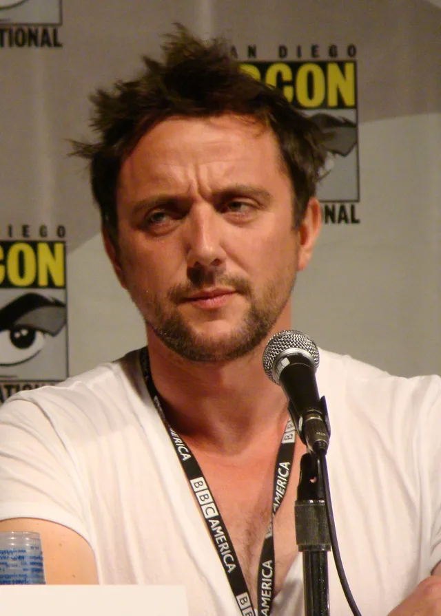 Peter Serafinowicz with tousled dark hair and stubble, seated at a microphone at a convention panel, wearing a white t-shirt.