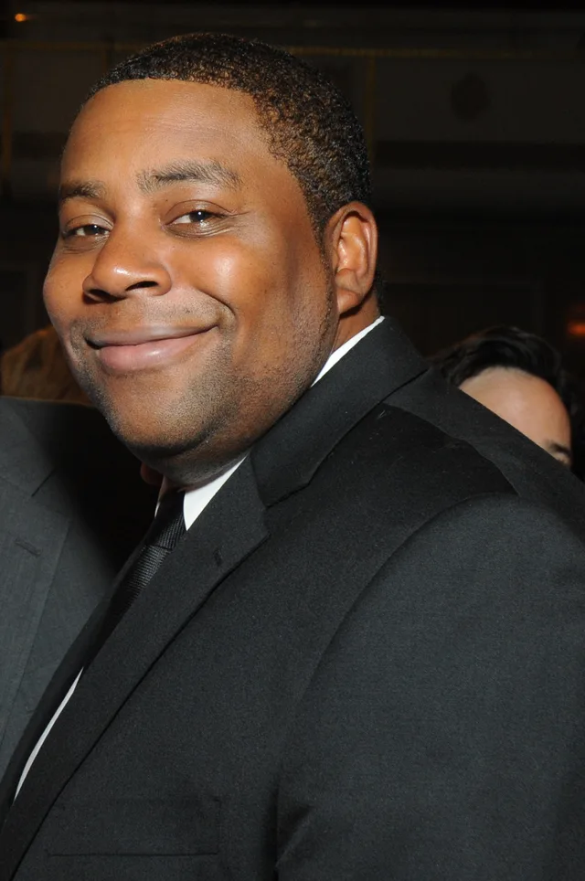 Colour photograph of a smiling man with short dark hair in a black suit, white shirt and dark tie, indoors at an evening event.