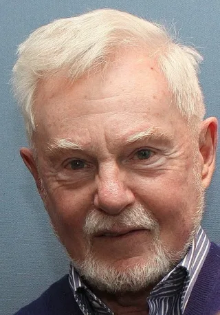 Close portrait of Derek Jacobi with white hair and a short white beard, in a purple jumper and striped shirt.