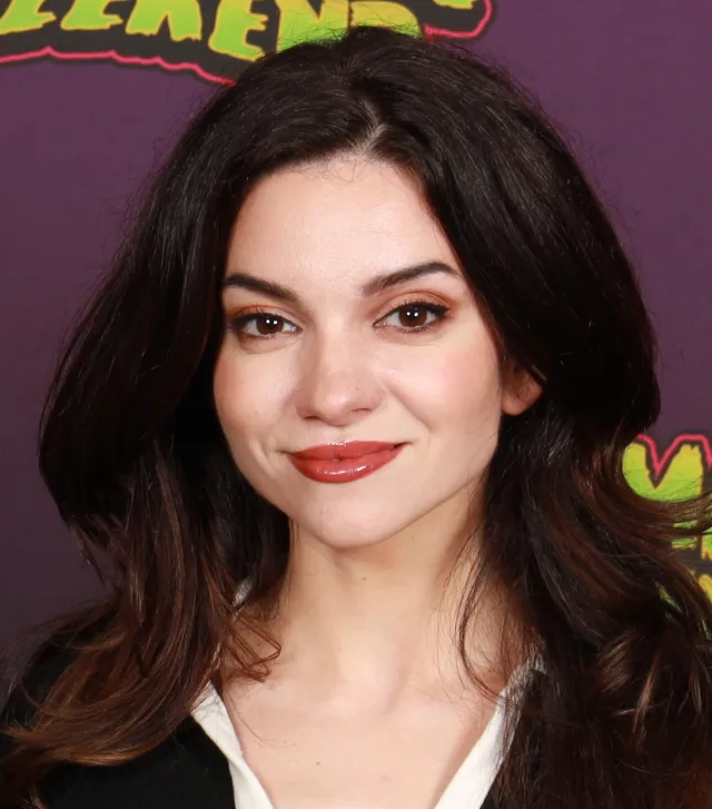 Lauren LaVera with long dark wavy hair and red lipstick, smiling slightly in front of a purple event backdrop.