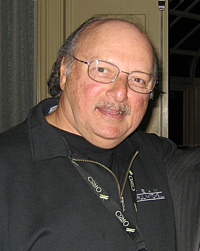 Portrait of a bald older man with a grey moustache and wire-framed glasses, smiling slightly, in a dark zip-neck top with a lanyard.