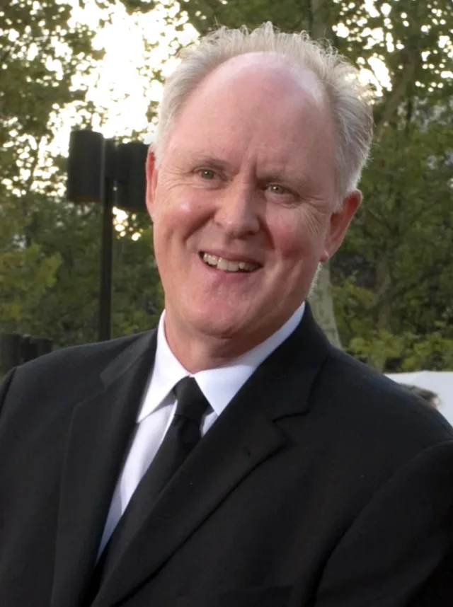 John Lithgow smiling outdoors in a black suit, white shirt and black tie, with trees and evening light behind him.