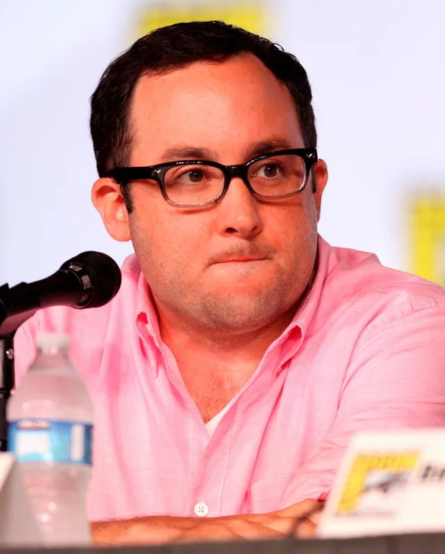 A man with dark hair and black-framed glasses in a pink shirt, seated at a convention panel table with a microphone.