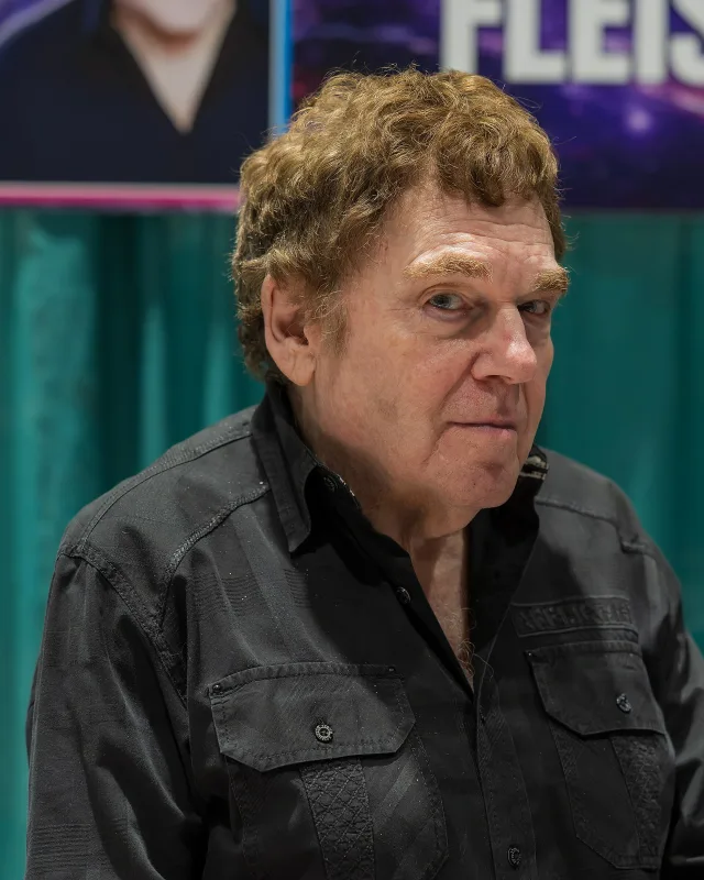 Older man with curly light-brown hair and heavy eyebrows, in a black button shirt, looking at the camera in front of a teal curtain.