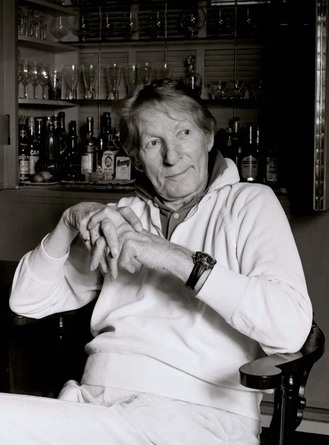 Black and white photograph of an older man in a white sweatshirt sitting in a chair with hands clasped, a shelf of glasses and bottles behind him.
