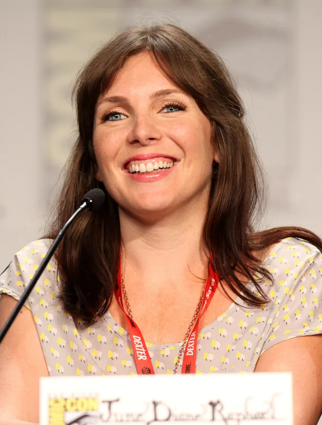 Smiling woman with shoulder-length brown hair and a fringe, in a patterned top with a red lanyard, seated at a panel microphone.