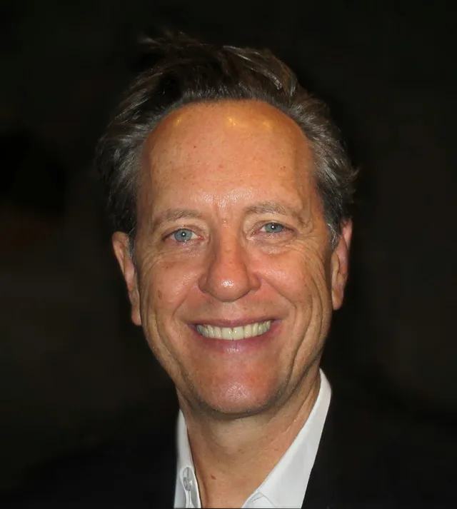 Head and shoulders photograph of a smiling man with swept-back grey-brown hair and blue eyes, wearing a dark jacket over a white shirt against a dark background.