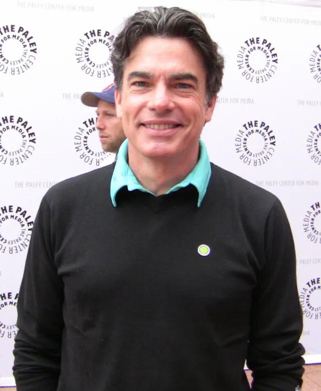 Smiling man with dark hair in a black jumper and teal collar, standing in front of an event backdrop.