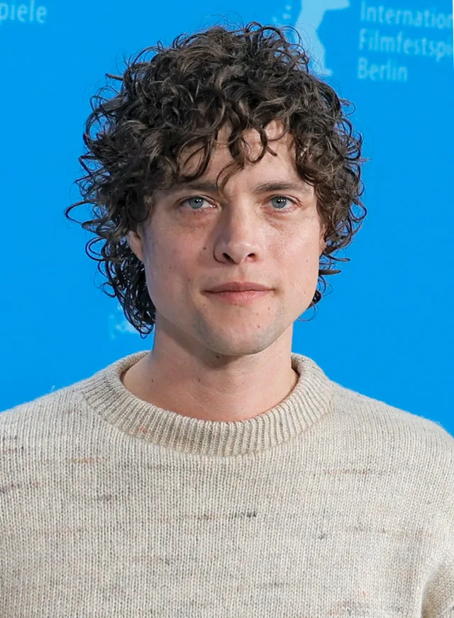Young man with curly dark hair and light eyes in a cream knitted jumper against a blue film festival backdrop