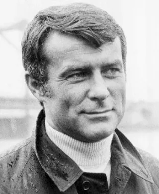 Black and white head and shoulders photograph of Robert Conrad in a rain-spotted jacket and polo neck, looking off to one side with a slight smile.