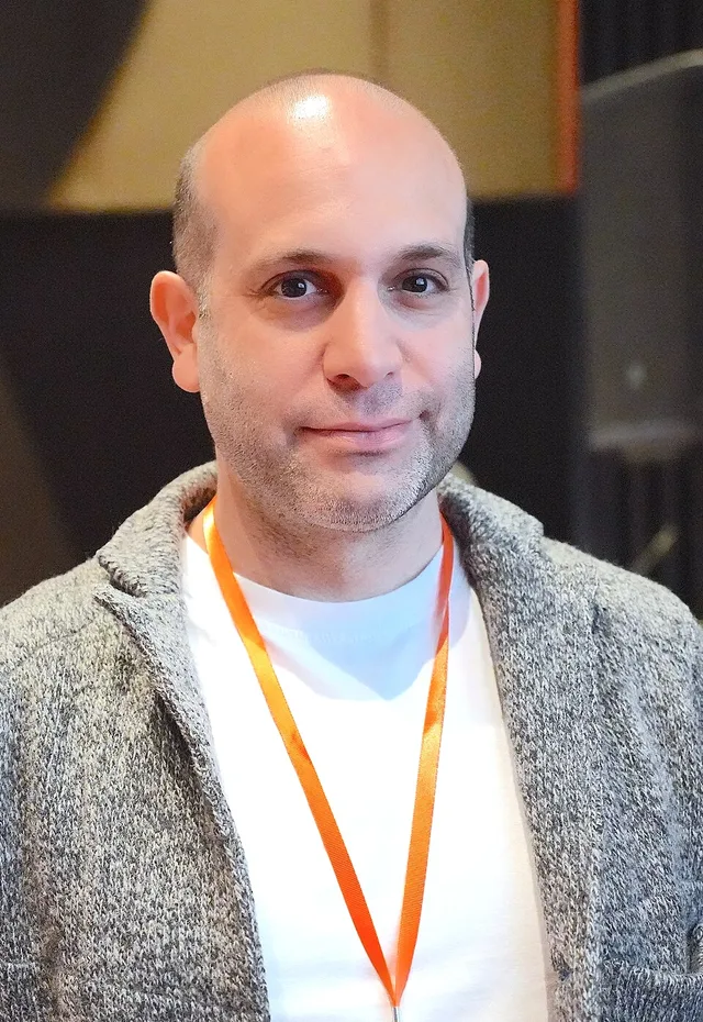 Bald man with light stubble in a grey knit cardigan over a white T-shirt, wearing an orange lanyard indoors.