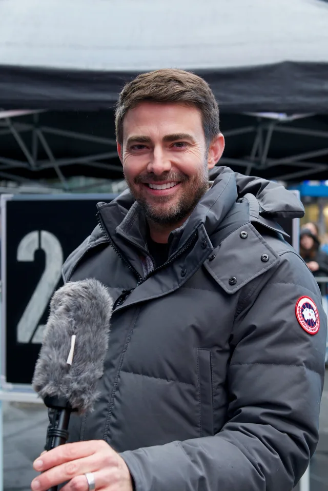 Smiling man with short brown hair and stubble in a grey padded winter jacket, holding a furry microphone outdoors.