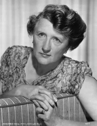 Black and white studio portrait of an older woman with short wavy hair, leaning on the back of a striped chair with her hands clasped.