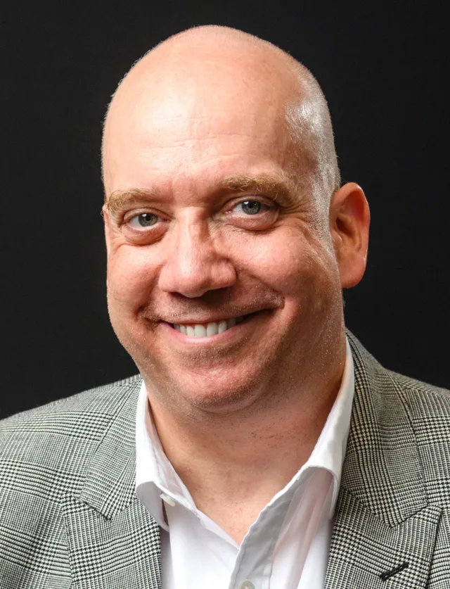Head and shoulders portrait of a smiling bald man in a grey houndstooth jacket and white open-collar shirt against a dark background.