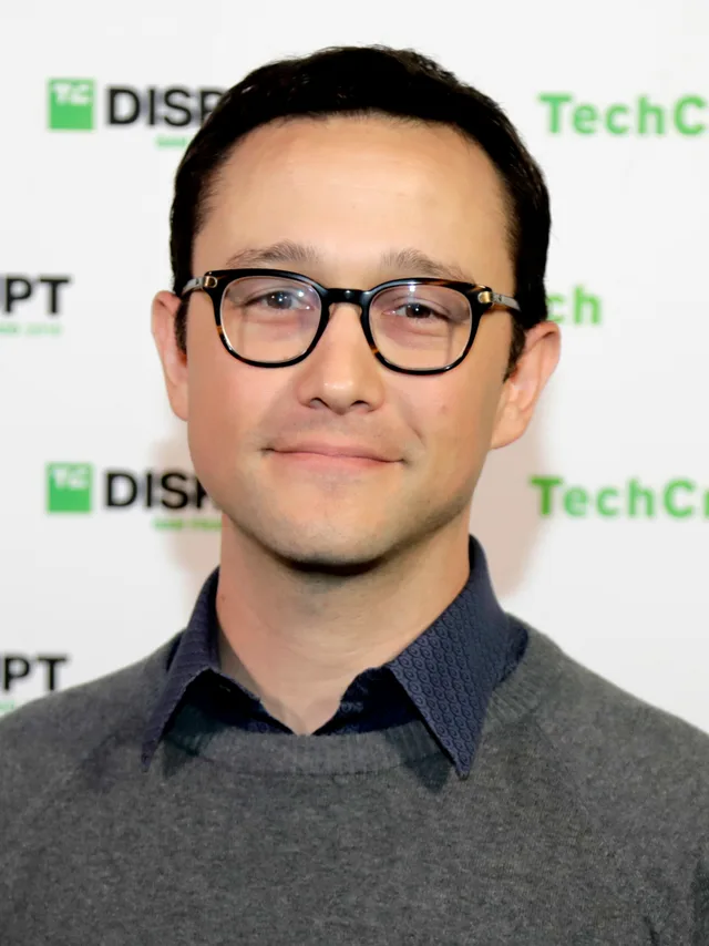 Portrait of a smiling man with short dark hair and dark-rimmed glasses, wearing a grey jumper over a blue patterned collar, in front of a conference backdrop.