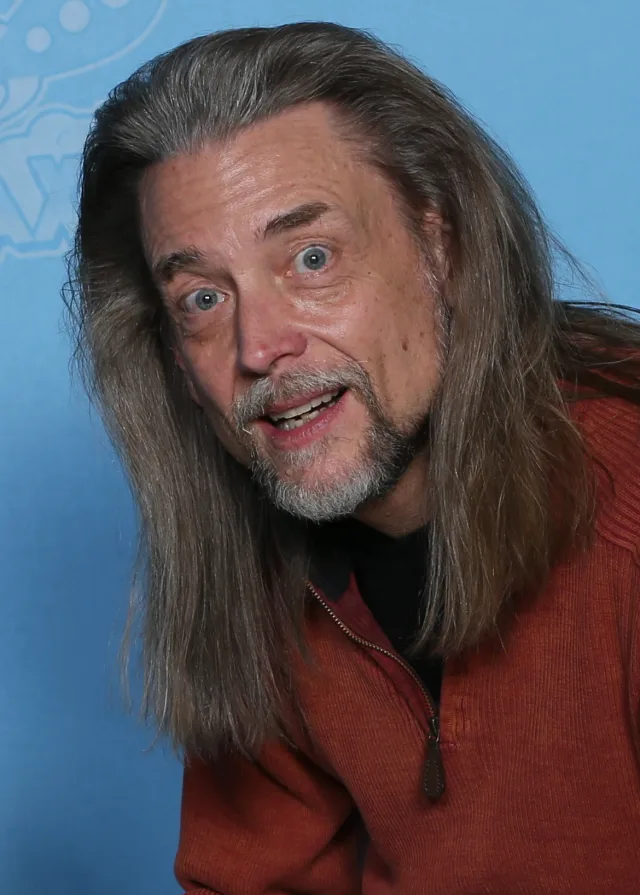 Steve Whitmire with long grey hair and a goatee, leaning toward the camera with wide eyes, in an orange zip-neck jumper against a blue backdrop.
