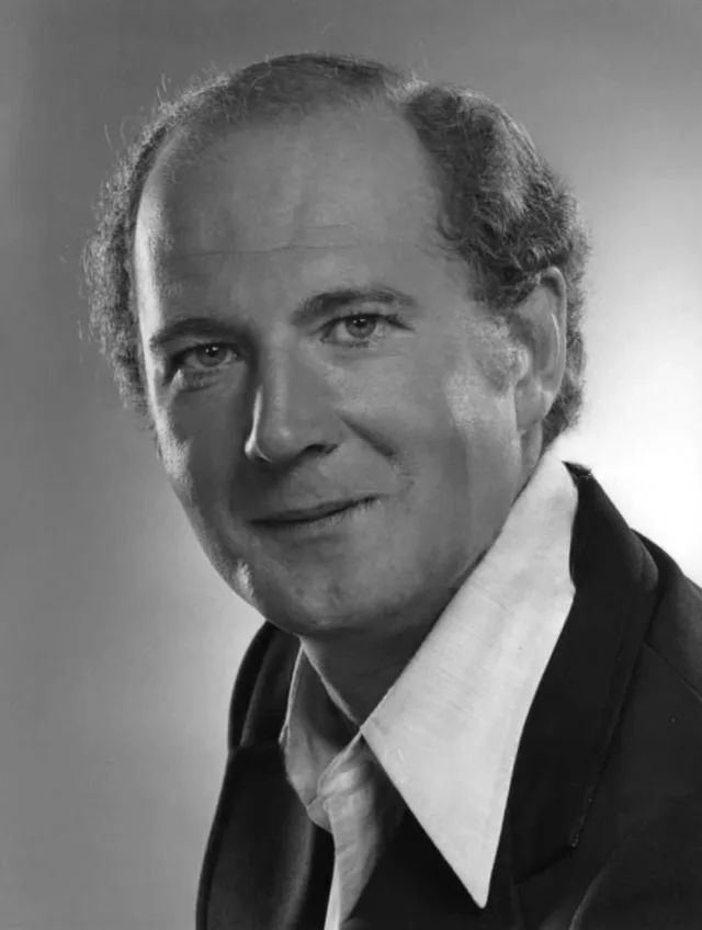 Black and white studio portrait of a smiling man with wavy hair, wearing a dark jacket over an open white collar.