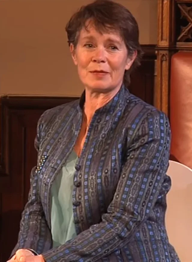Celia Imrie seated on stage in a patterned blue jacket over a pale green top, with short dark hair and a slight smile.