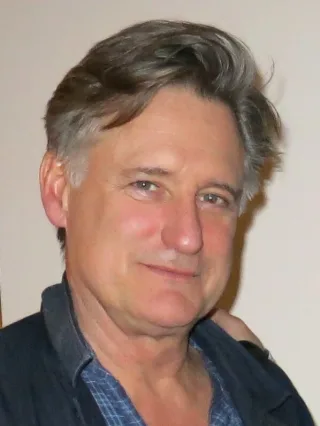 Close photograph of a man with swept grey hair, wearing a dark jacket over a blue checked shirt.