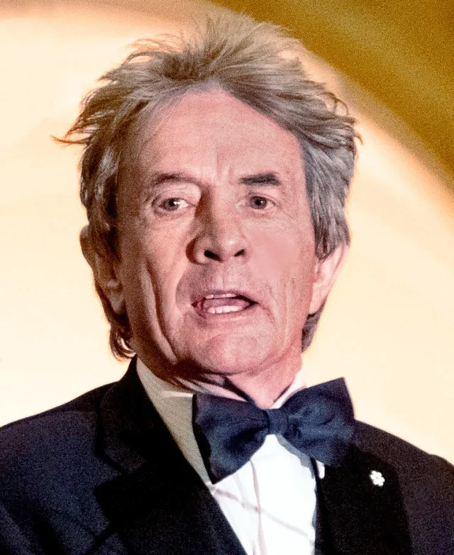 Martin Short mid-sentence, with swept-up grey hair, a black dinner jacket and bow tie, against a warm gold background.