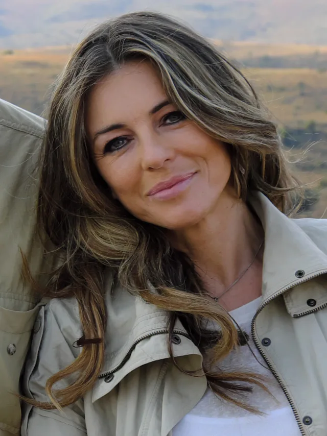 Elizabeth Hurley with long highlighted hair, smiling slightly, in a stone-coloured jacket against open countryside