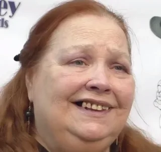 Tight headshot of a smiling older woman with long red hair and dark beaded earrings, in front of a white event backdrop.