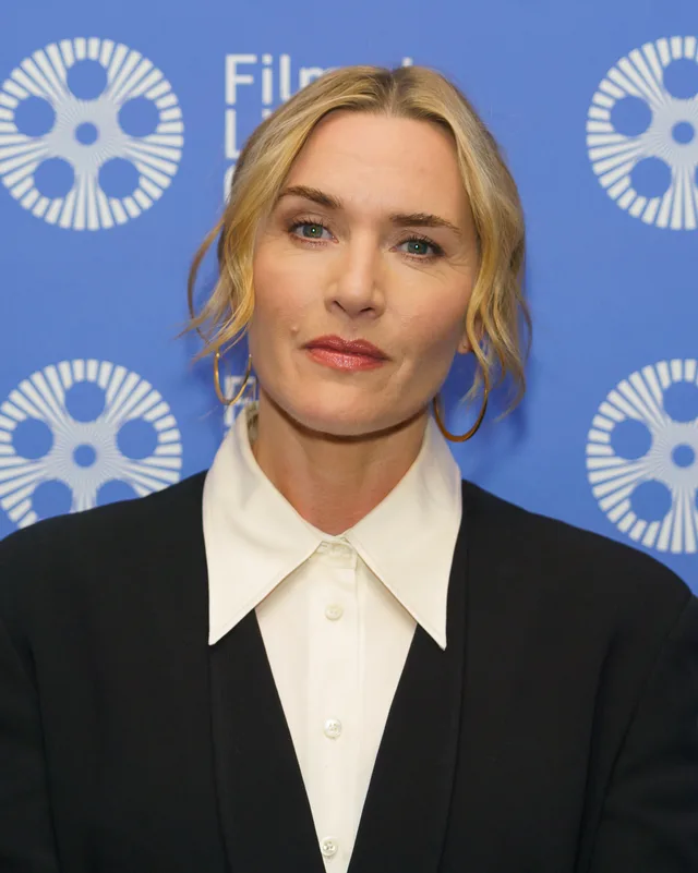 Head and shoulders photograph of Kate Winslet with wavy blonde hair and gold hoop earrings, wearing a black jacket over a white collared shirt, in front of a blue backdrop.