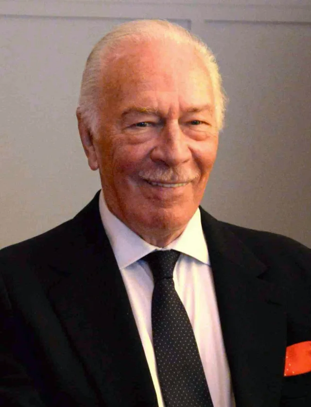 Smiling older man with swept-back white hair, wearing a dark suit, white shirt, dotted tie and an orange pocket square