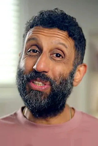 Close portrait of a man with short dark curly hair and a full greying beard, wearing a dusty pink top and speaking to the camera indoors.