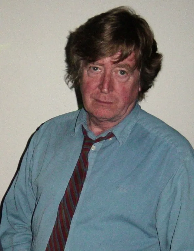 A middle-aged man with tousled brown hair in a blue shirt and a striped red and blue tie, looking at the camera against a plain wall.