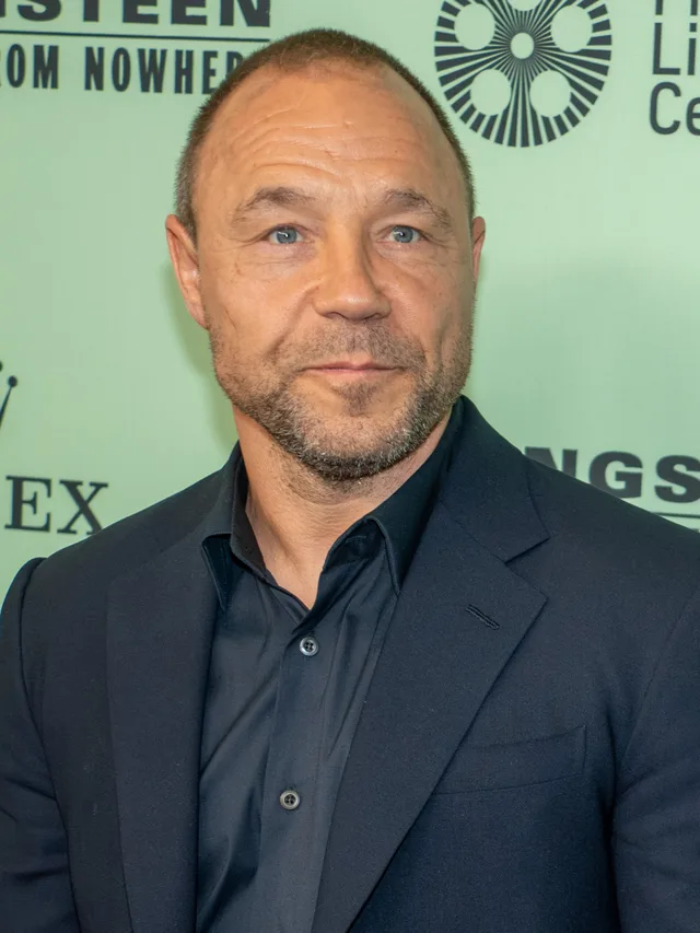 Portrait of a man with a shaved head and greying stubble in a dark navy jacket and shirt, standing in front of a pale green event backdrop.