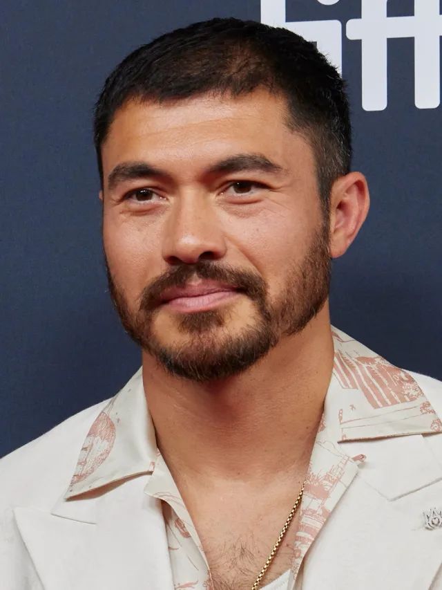Close photograph of a bearded man with short dark hair in a cream jacket and patterned shirt with a gold chain, against a navy backdrop.