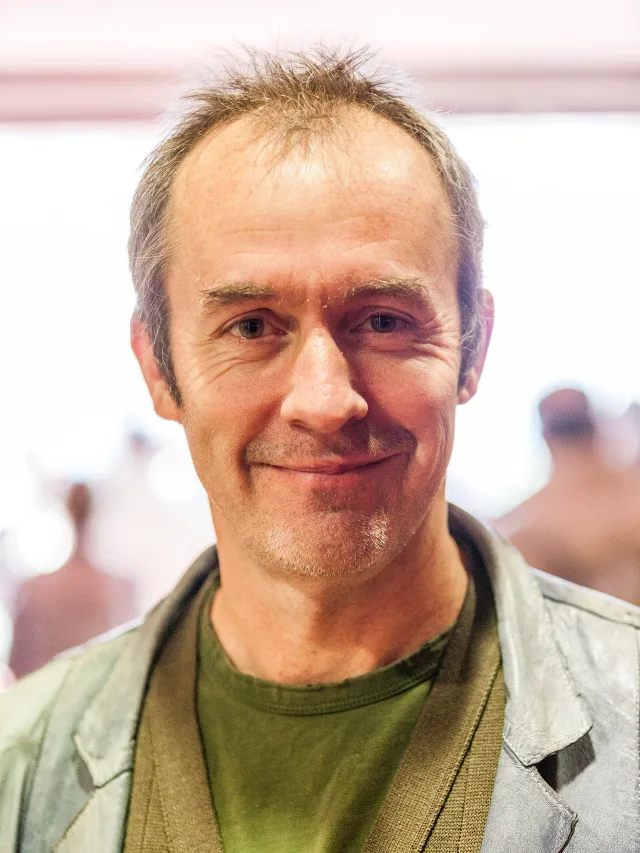 Head and shoulders photograph of Stephen Dillane, smiling slightly, with short greying hair, wearing a grey jacket over a green top.
