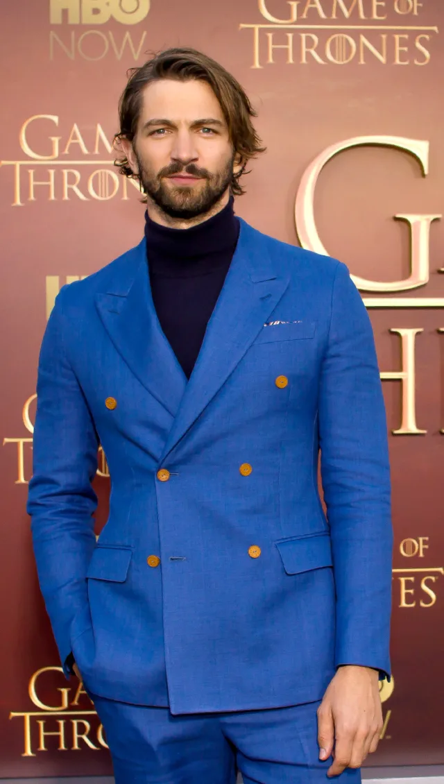 Bearded man with swept-back brown hair in a bright blue double-breasted suit and navy polo neck, standing with one hand in a pocket.