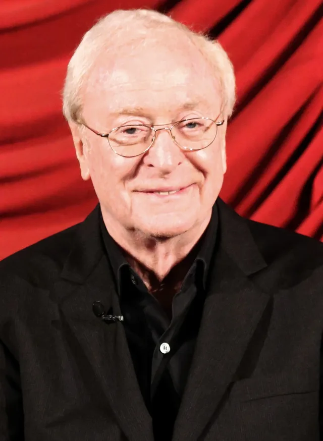 Michael Caine smiling gently at the camera, wearing thin-rimmed glasses and a dark jacket in front of a red curtain.