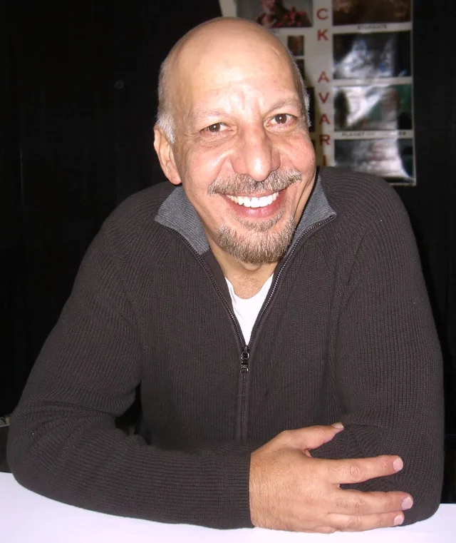 A smiling bald man with a grey goatee in a dark zip-up jumper, seated at a table with his hands folded in front of him.