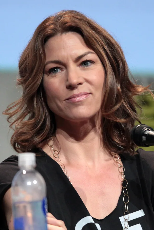 Portrait of a woman with wavy shoulder-length brown hair and a gold chain necklace, seated at a panel microphone.