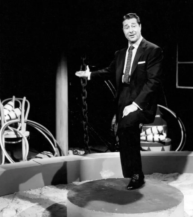 Black and white photograph of a man in a dark suit and patterned tie gesturing with one hand, standing on a small stage set.